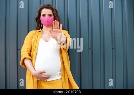 Pregnant woman in a protective mask and yellow clothes on a blue wall background. The concept of social distance.