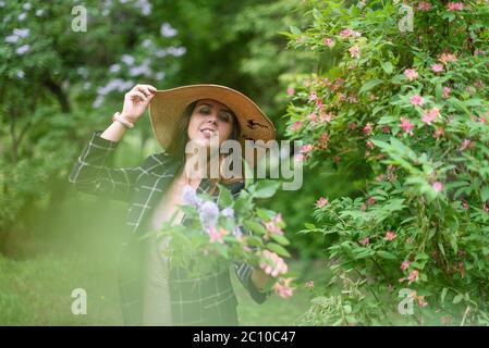 Girl in straw summer hat in pink dress and dark jacket holds her hat and bouquet against a background of blooming trees Stock Photo