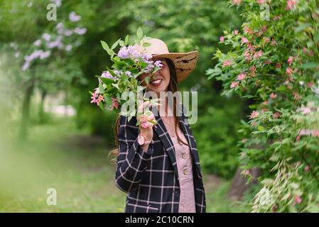Girl in straw summer hat, pink dress and dark jacket hide her face with bouquet against a background of blooming trees Stock Photo