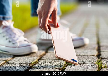 Woman hand Picking broken phone from cobblestoned pavement after drop, bottom view, close up. Smartphone with broken glass on the back cover. Stock Photo