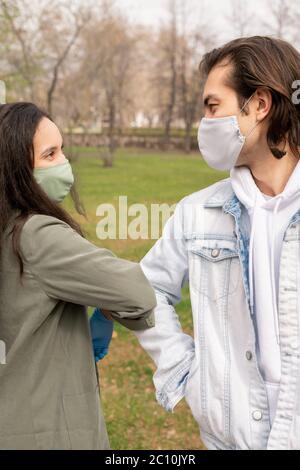 Beautiful couple in cloth masks touching elbows instead of kiss while meeting in park Stock Photo