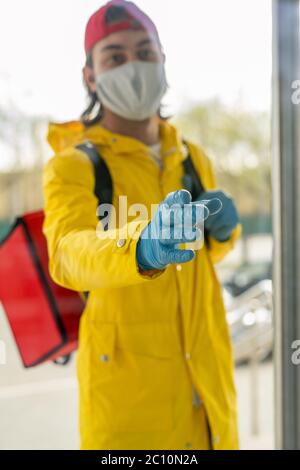 Delivery man in mask and latex gloves wearing thermo bag behind back and reaching hand to open door Stock Photo