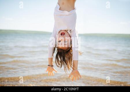 Little hiperemotional school age girl hanging upside down. On a lake shoreside. Stock Photo