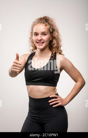Pretty young fit sportswoman in black tracksuit showing thumb up and expressing success in physical trainings while looking at you Stock Photo