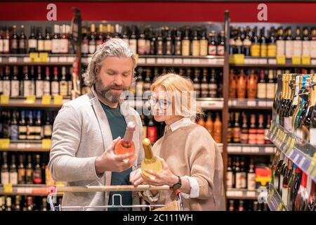 Aged bearded man with bottle of rose wine and his blond wife reading information on label while choosing alcoholic drinks Stock Photo