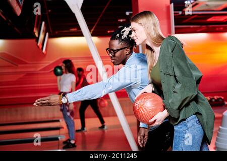 Young man of African ethnicity pointing at bowling track while explaining his girlfriend how to throw ball to strike all skittles Stock Photo