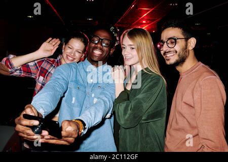 Four young cheerful intercultural friends looking in smartphone camera while African guy making selfie in dark room of leisure center Stock Photo
