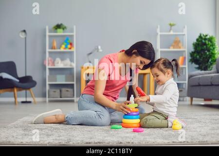 Young mother and her daughter engaged in interesting game on floor at home. Parent and kid playing with pyramid indoors Stock Photo