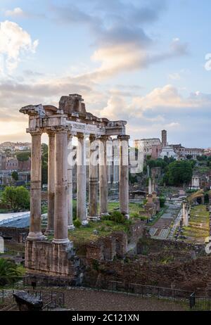 Columns of the ancient Temple of Saturn at sunrise, in the Roman Forum, Rome, Italy, with ruins of Basilica Giulia,  temples and the Colosseum amphith Stock Photo