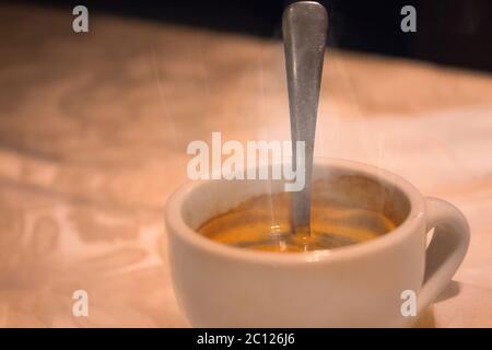 Smoke emitted from a cup with hot coffee and teaspoon in it. It placed on a tablecloth on a table. Close up with selective focus. Stock Photo