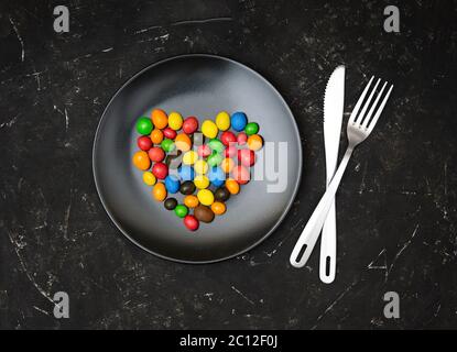 Multicolored candy dragees arranged in shape of a heart on a matte black plate, knife and fork on a textured black background. Flat lay, top view. Var Stock Photo