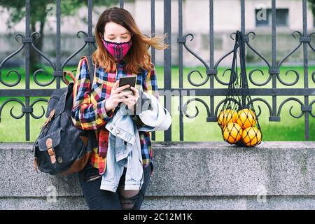 Young woman wearing a plaid handmade face mask and colorful shirt with mesh bag full of oranges and a backpack standing on the street. Stock Photo