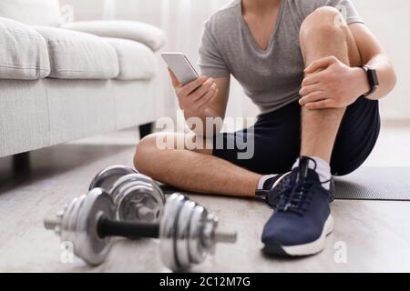Power training. Guy with fitness tracker and smartphone sits on floor with dumbbells Stock Photo