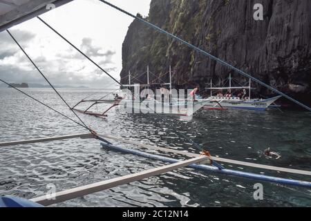 Tourists on traditional Philippino fisherman boats crossing the waters near grey and green limestone cliffs on a cloudy day of El Nido at Palawan Isla Stock Photo