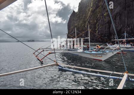 Tourists on traditional Philippino fisherman boats crossing the waters near grey limestone cliffs on a cloudy day of El Nido at Palawan Island the Phi Stock Photo