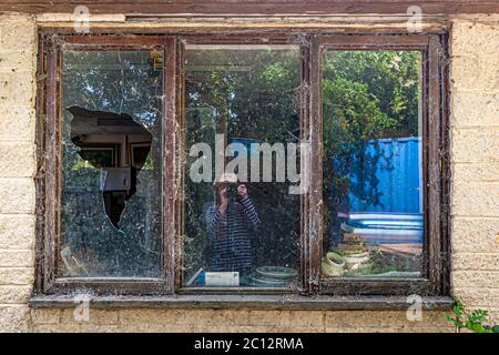 A photographer is reflected in the middle pane of a three-part sash window on an abandoned house, its left pane shattered and covered in cobwebs, in Lechlade, Gloucestershire, England. Stock Photo