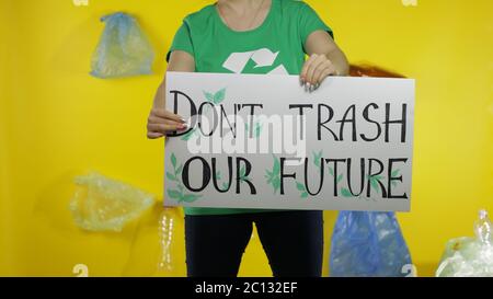 Unrecognizable woman volunteer in t-shirt with recycle logo holding protesting message poster Don't Trash Our Future. Background with cellophane bags, bottles. Environment trash plastic pollution Stock Photo