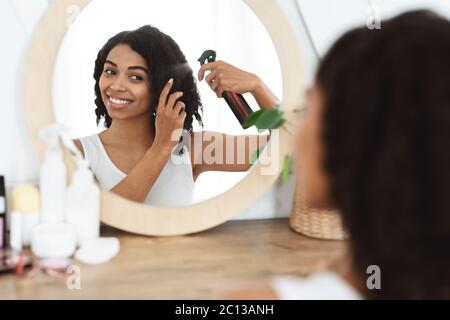 Hairstyling. Beautiful Black Woman Using Hair Spray For Easy Combing Near Mirror Stock Photo