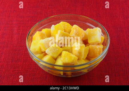Frozen mango fruit chunk cubes with ice crystals in glass ingredient bowl on red tablecloth Stock Photo