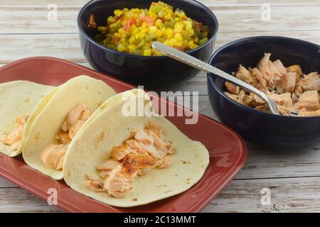 Making salmon tacos by putting salmon in tortillas on plate with two filling bowls of baked salmon fillet pieces and mixture of corn, tomatoes, green Stock Photo