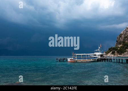 Transport Boats are docked next to wooden pier and a body of water, with stormy sky and mountain covered with trees at the background, High quality ph Stock Photo