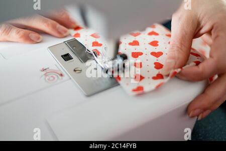 Woman hands using the sewing machine to sew the face mask during the coronavirus pandemia. Homemade protective mask and pieces of cloth on a gray back Stock Photo