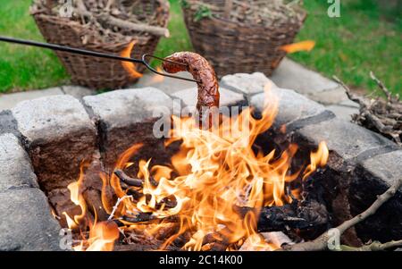 Toasting sausages on campfire in nature. Dinner or lunch in nature Tasty sausage stabbed on an iron fork. Closeup shot toasted over an fire. Stock Photo