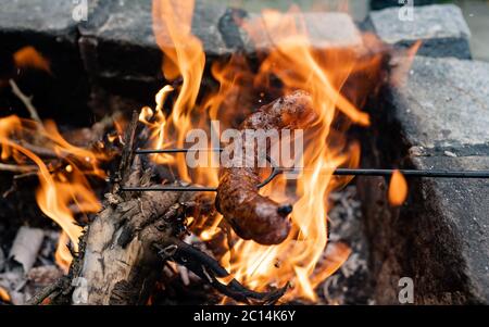 Sausage stabbed on an iron fork and toasted over on campfire in nature. Baking sausages. Closeup shot on fire and on roasted sausage. Stock Photo