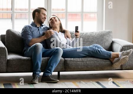 Happy young couple hugging, relaxing on couch, using smartphone Stock Photo