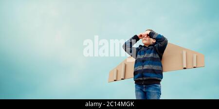 Child boy playing with paper wings airplane and dreaming of becoming a pilot. Kid on the blue sky background outdoors. The concept of teamwork Stock Photo