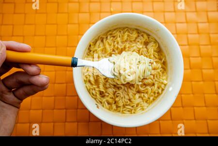 Hand of a man holding a fork with cooked instant noodles from ceramic bowl on orange background. Top view. Cheap food Stock Photo