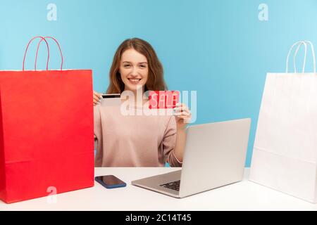 Happy woman buyer surrounded by packages smiling and holding credit card with Sale inscription, making purchases online on laptop using shopping loan. Stock Photo