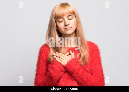 Cute young blonde woman in fluffy red sweater keeps both palms on chest near heart, closed eyes, has charming smile, expresses good emotions, isolated Stock Photo