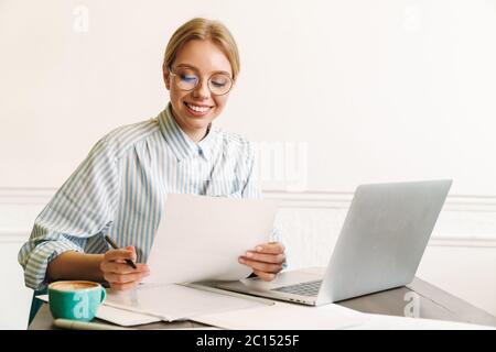 Photo of smiling blonde woman architect in eyeglasses working with laptop while designing draft at workplace Stock Photo