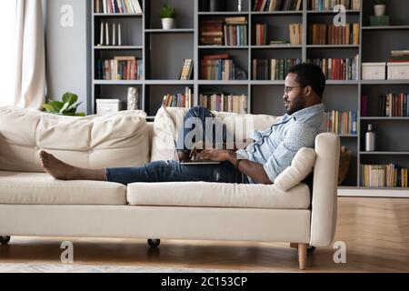 African guy lying on sofa typing message using computer Stock Photo
