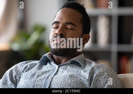 Close up African young man sit on couch closed eyes resting reduces fatigue Stock Photo