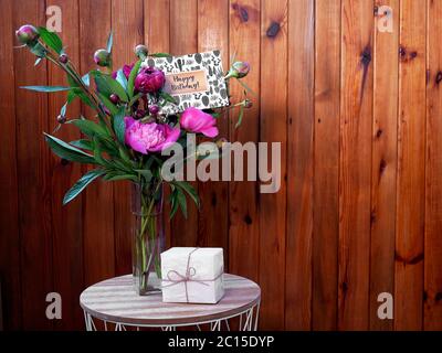 Pink flowers peonies in a vase on a wooden background with a small white gift and a happy birthday card. The photo was taken with natural daylight. Stock Photo