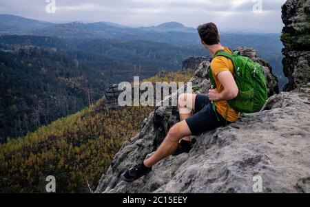 Fearless hiker man in sportswear sitting on cliff edge. Sandstone rock, forest hilly landscape and green view. Nature park Alone with nature. Stock Photo