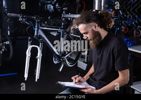 Stylish bicycle mechanic making notes in clipboard in workshop. theme small business selling, repair and service bicycles. Technician Doing Stock Photo
