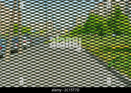 Dark iron lattice with rhombic cells through which the street is visible on a bright sunny day. Stock Photo