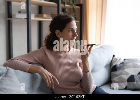 Smiling woman recording voice message, holding phone, sitting on couch Stock Photo