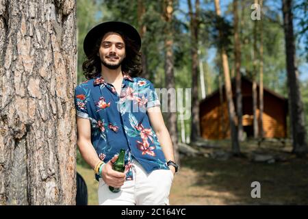 Portrait of positive hippie guy in flower shirt and hat leaning on tree and holding beer bottle in forest Stock Photo