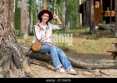 Portrait of smiling pretty young woman in jeans and hat sitting on root of tree while resting in recreation center Stock Photo