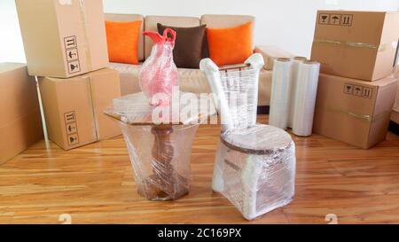 Moving scene with a chair, a vase on a table packed in plastic inside a room with a sofa, plastic rolls and closed cardboard boxes on white background Stock Photo