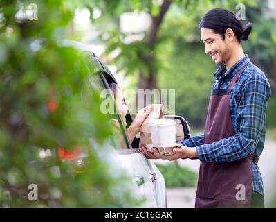 Waiter giving hot coffee cup with disposable tray and bakery bag through car window to customer at drive thru service station. Drive thru is popular s Stock Photo