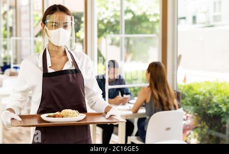 Portrait attractive asian waitress wear face mask and face shield holding food tray to serving meal to customer with custome in background. New normal Stock Photo