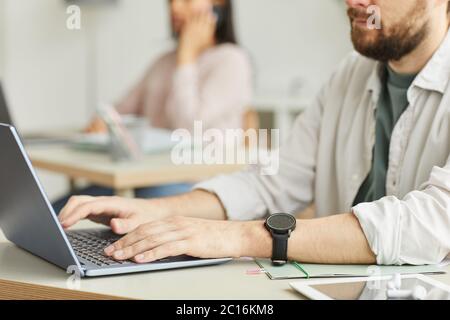 Side view close up of unrecognizable man using laptop while working in office space with colleagues in background Stock Photo