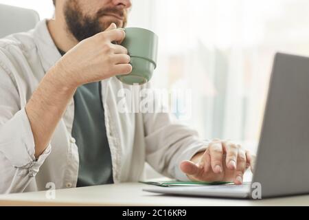 Cropped portrait of bearded adult man drinking coffee while using laptop at home office, copy space Stock Photo