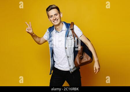 A young man in a denim outfit is holding a travel bag and showing a peace gesture, isolated over a yellow background. Young traveler. Stock Photo