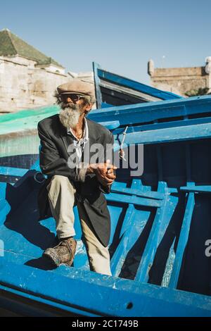 ESSAOUIRA, MOROCCO - SEPTEMBER 10, 2019: Old senior fisherman on blue wooden boat in Essaouira harbor. Stylish look, hat, beard, sunglasses and jacket. Moroccan people. Stock Photo